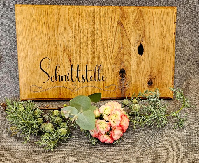 Brotzeitbrett Holztafel mit dem Schriftzug "Schmidtstelle" und einer Blumenarrangement.